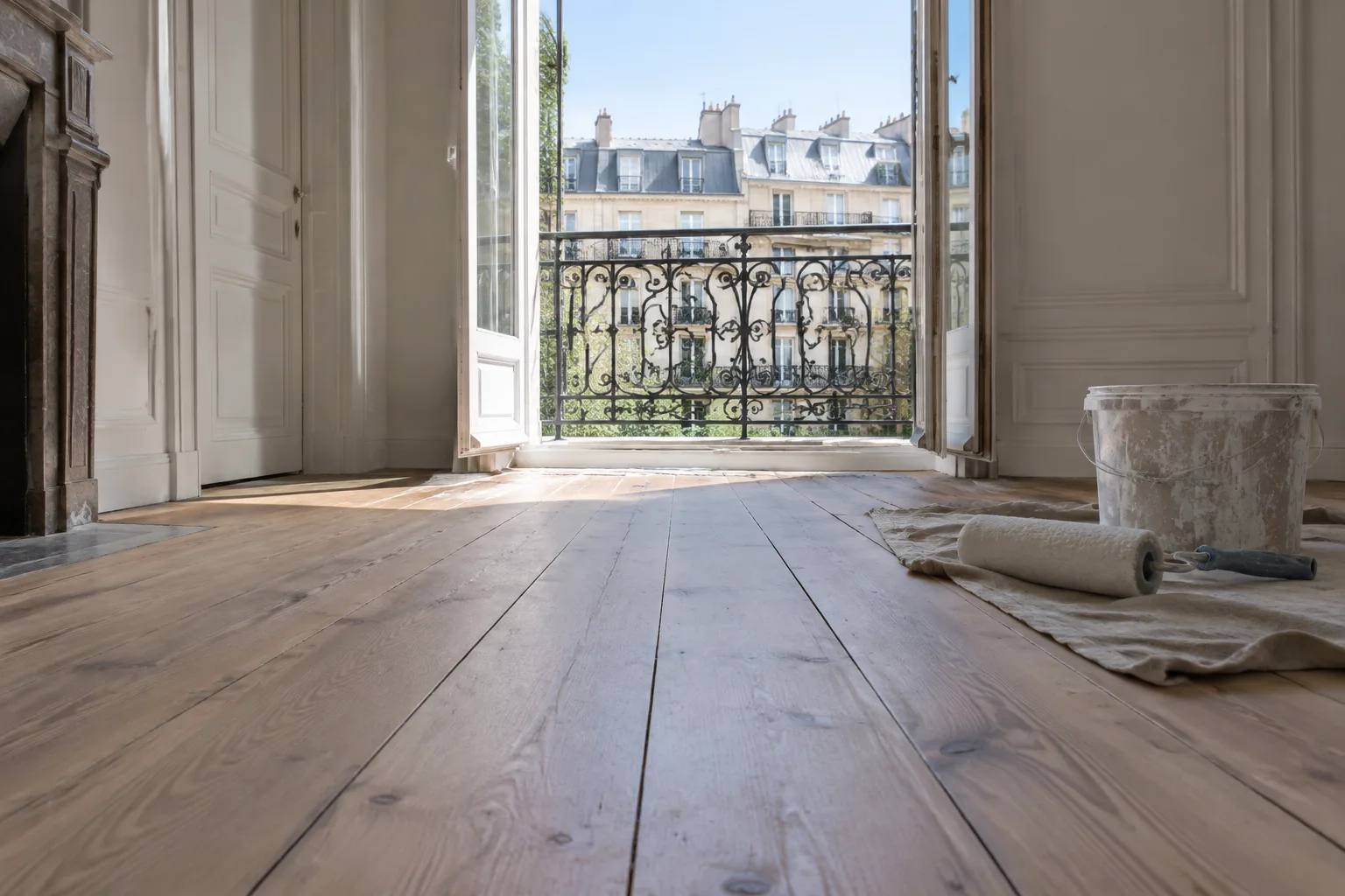 Parquet poncé d'un salon parisien menant à un balcon en fer forgé, avec pot de peinture au sol