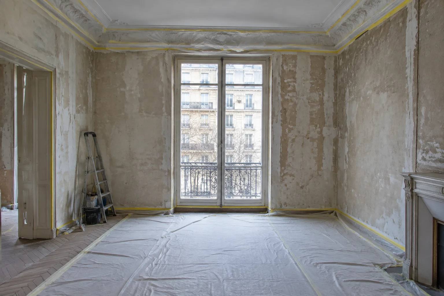 Stripped Haussmannian room mid-renovation with masked mouldings, bare plaster walls and covered parquet by a tall window